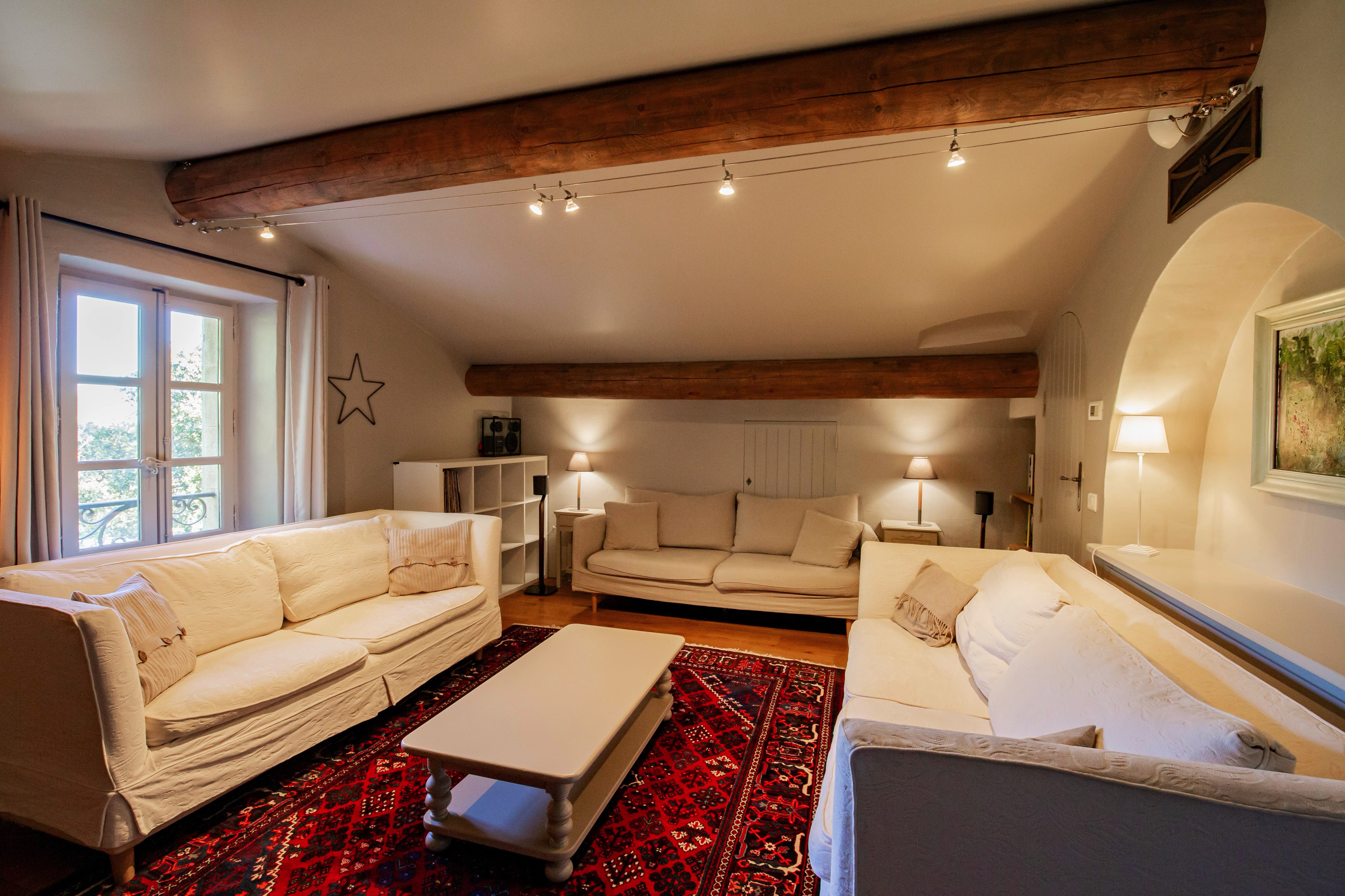 Cozy living room featuring white sofas, a central coffee table, red oriental carpet, and wooden ceiling beams, with soft lighting and a window allowing natural light.